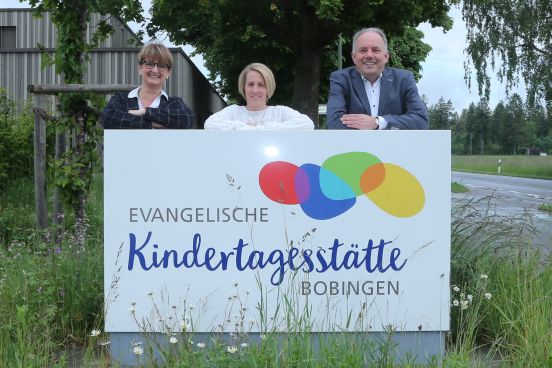 Die Leiterinnen der Kita und der Pfarrer stehen lächelnd vor dem Schild der Kindertagesstätte.